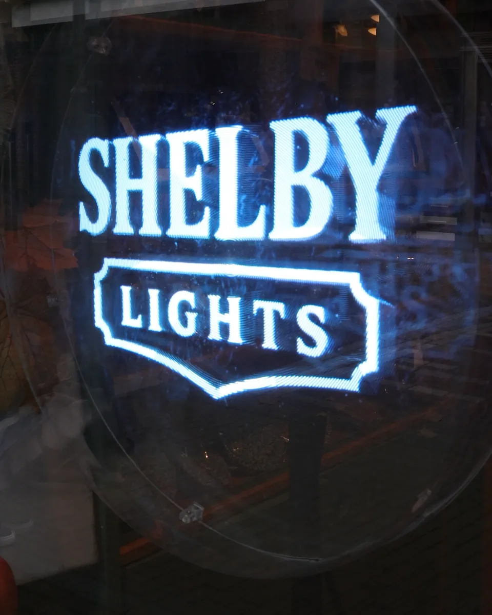 The Shelby Lights logo as a hologram behind a shop window, with the street reflected in the glass